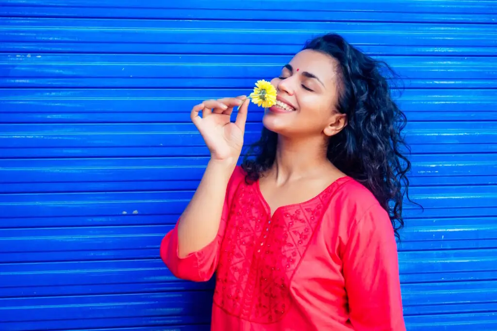 vAttractive beautiful happy young latin hispanic woman with teak bindi on forehead smiling on blue wall street background holding fresh flover by John FRANCEY